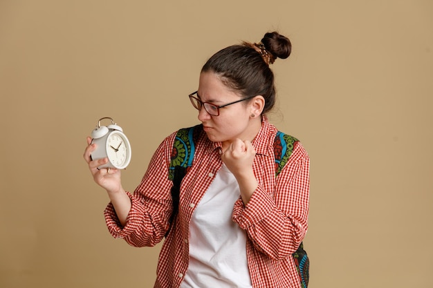 茶色の背景の上に立って怒りで拳を握りしめる怒った顔でそれを見て目覚まし時計を保持しているバックパックと眼鏡をかけているカジュアルな服を着た学生の若い女性