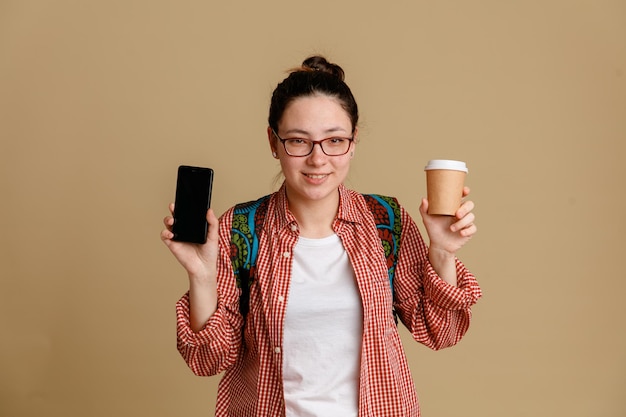 コーヒー カップと携帯電話を保持しているバックパックとメガネを身に着けているカジュアルな服を着た学生の若い女性は、カメラを見て幸せで肯定的な笑みを浮かべて、茶色の背景の上に自信を持って立っています。