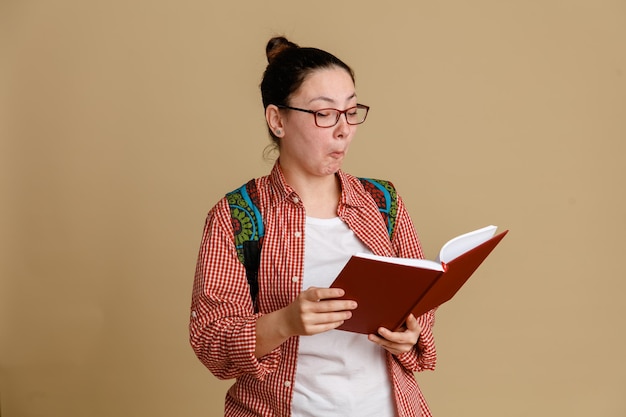 Студентка в повседневной одежде в очках с рюкзаком, держащая блокнот, выглядит заинтригованной, читая что-то стоящее на коричневом фоне