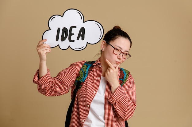 Student young woman in casual clothes wearing glasses with backpack holding speech bubble with word idea looking at camera with pensive expression standing over brown background