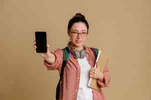 Free photo student young woman in casual clothes wearing glasses with headphones and backpack holding notebook and mobile phone showing at camera smiling confident standing over brown background