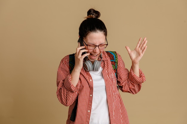 ヘッドフォンとバックパックを身に着けているメガネを着たカジュアルな服を着た学生の若い女性が携帯電話で話し、怒って怒って茶色の背景の上に立っています