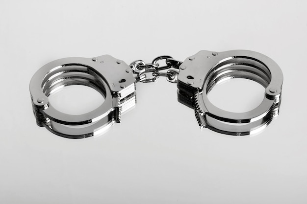 Студийный крупный план запертой пары наручников типа хиатт, отражающихся на блестящей поверхности как символ тюремного заключения и преступности