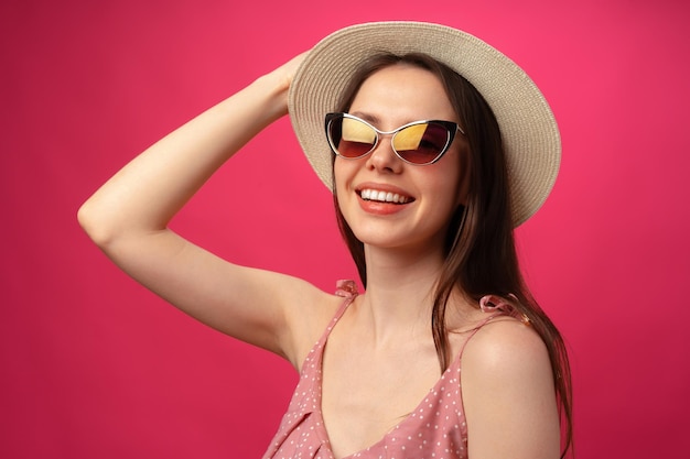 Studio fashion portrait of a young attractive woman in hat and glasses against pink backgorund