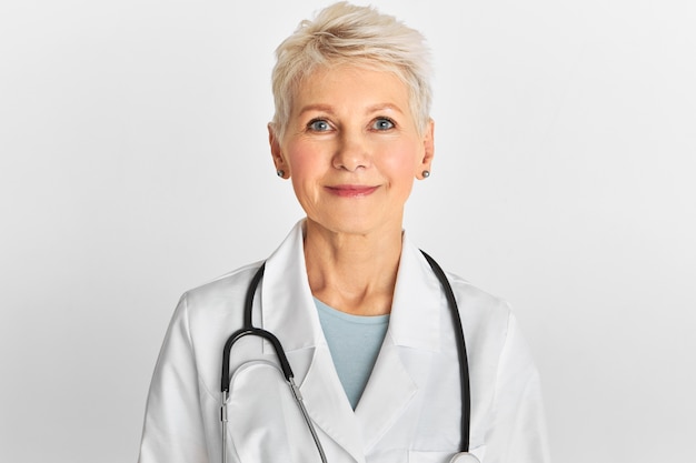 Studio image of confident attractive middle aged female doctor with short dyed hairstyle posing isolated wearing white coat and stethoscope.