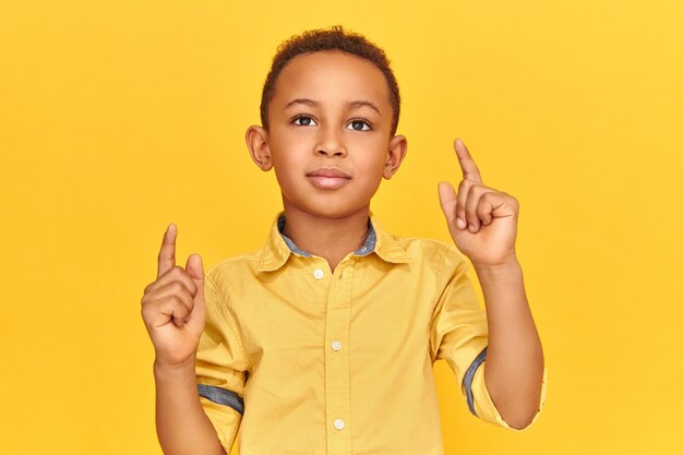 자신감이 멋진 어두운 피부 어린 소년의 스튜디오 이미지 복사 공간 f를 나타내는 앞쪽 손가락을 위쪽으로 가리키는 노란색 벽 배경에 고립 된 포즈