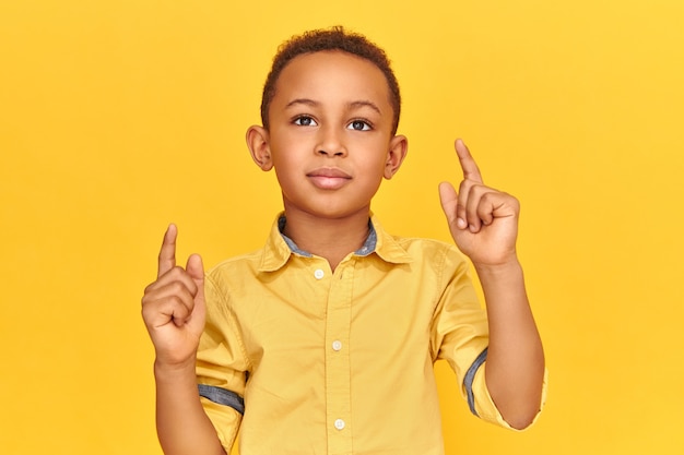 Studio image of confident cool dark skinned little boy posing isolated against yellow wall background pointing fore fingers upwards, indicating copy space f
