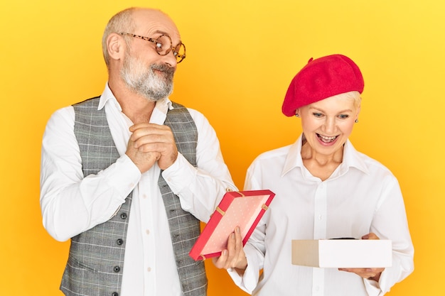 Studio image of emotional elegant male pensioner with gray beard and bald head looking with impatience, watching attractive woman opening box with present from him. Excited mature lady receiving gift