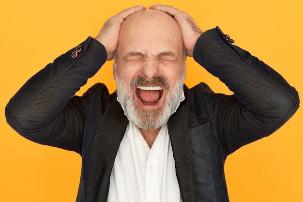 Free photo studio image of enraged devastated elderly businssman in formal clothes closing eyes and screaming out loud, loosing temper, keeping hands on his bald head, stressed because of business failure