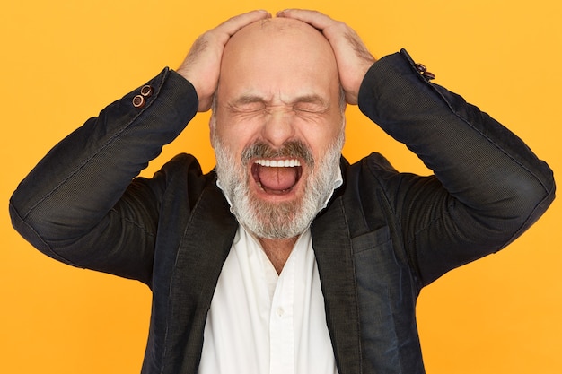 Studio image of enraged devastated elderly businssman in formal clothes closing eyes and screaming out loud, loosing temper, keeping hands on his bald head, stressed because of business failure