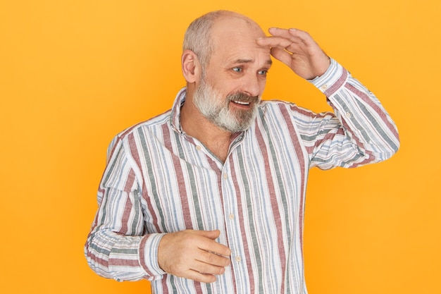 Free photo studio image of frustrated puzzled senior bald unshaven man holding palm on forehead to protect eyes from sun, looking into dustance or searching for something.