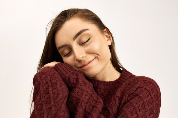 Studio image of gorgeous happy young dark haired woman closing eyes with pleasure, putting cheek on hands, wearing stylish warm sweater. Cute girl enjoying soft yarn on new jumper, smiling