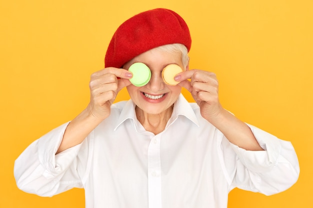 Studio immagine di gioiosa donna in pensione in camicia bianca e rosso bonner divertendosi tenendo due biscotti francesi rotondi sui suoi occhi.