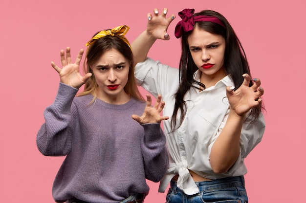 Studio image of two emotional attractive young women acting aggressive grimacing having wild fearsome facial expressions making gesture trying to scare you. Human emotions, feelings and reaction