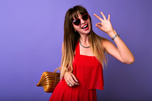 Studio lifestyle portrait of pretty brunette trendy woman wearing summer elegant red dress, Herat sunglasses, wooden bag.