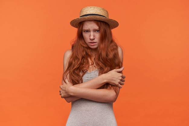 Studio photo of attractive young woman with wavy red lond hair hugging herself with hands, looking to camera fearfully and frowning her face, isolated over orange background