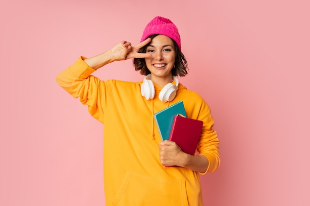 Studio photo of happy cute student with notebooks and earphones standing on pink. 