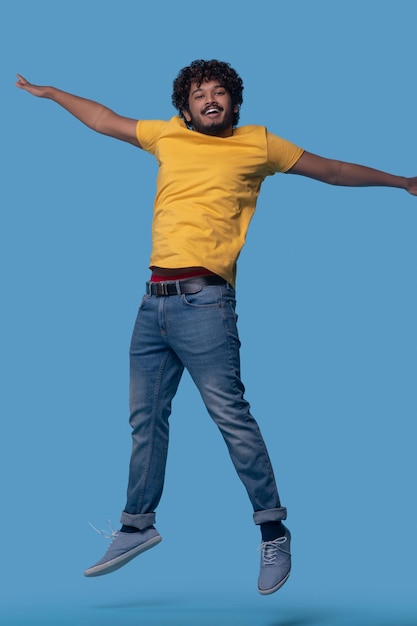 Studio photo of a smiling happy curly-haired Indian male leaping up against the blue background