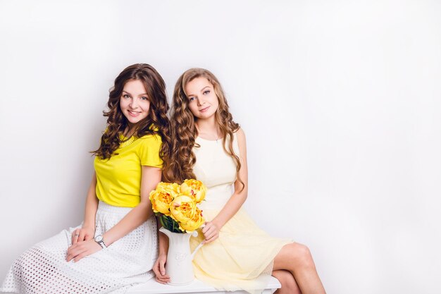 肩を並べて座っている2人の笑顔の女の子のスタジオ写真。ブルネットは白いスカートと黄色のTシャツを着ており、ブロンドの女の子は黄色のドレスを着て、黄色い花が付いた白い花瓶を持っています。