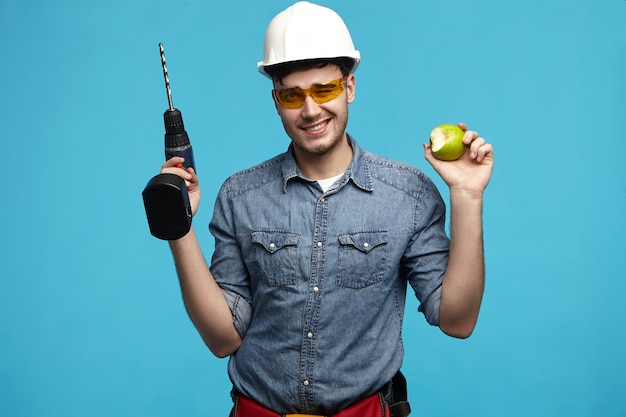 Студийное изображение трудолюбивого красивого молодого разнорабочего в каске и очках, позирующего
