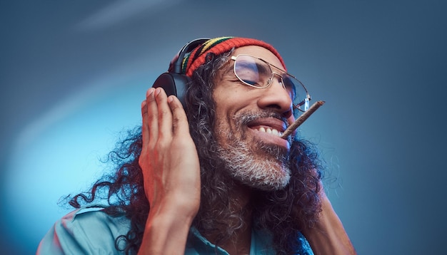 아프리카 Rastafarian 남성의 스튜디오 초상화는 헤드폰을 끼고 대마초를 피우는 음악을 즐깁니다. 파란색 배경에 고립.