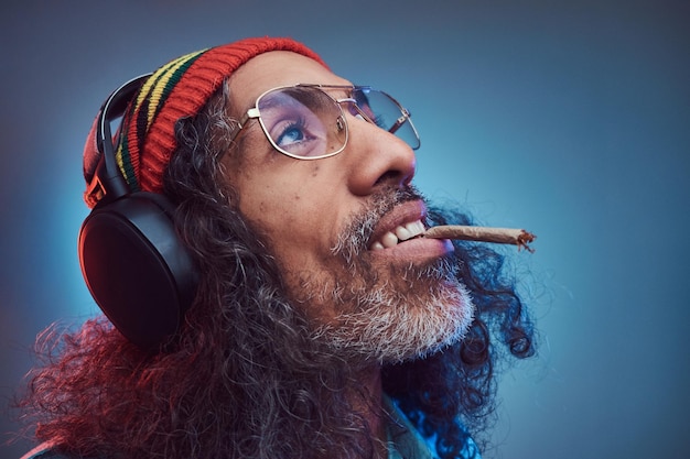 아프리카 Rastafarian 남성의 스튜디오 초상화는 헤드폰을 끼고 대마초를 피우는 음악을 즐깁니다. 파란색 배경에 고립.