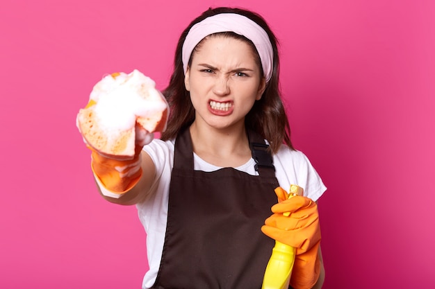 Studio portrait of angry young Caucasian woman dressed white t shirt and brown apron while cleaning up rooms, holding cleanser spray and sponge with foam in her hands, poses in photo studio.