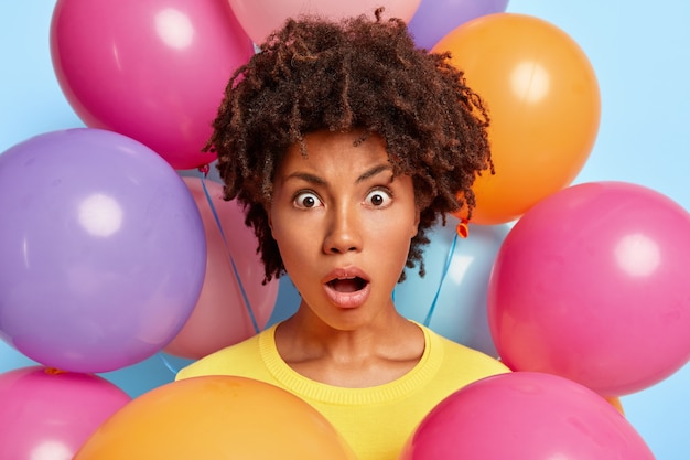 Студийный портрет изумленной молодой женщины, позирующей в окружении разноцветных шаров на день рождения