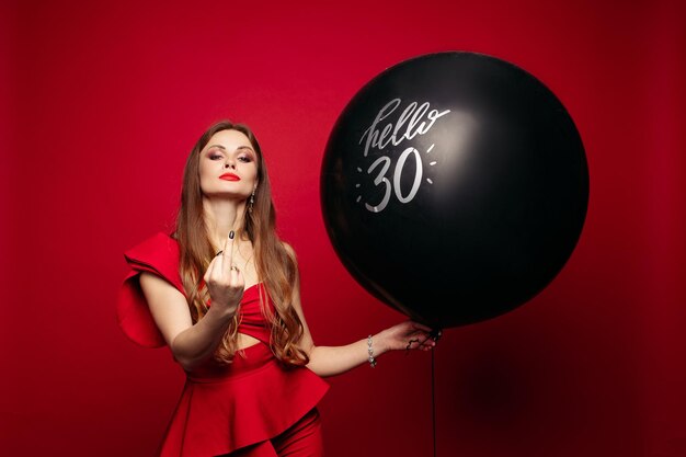검은 공기 풍선이 달린 세련된 빨간 드레스를 입은 매력적인 자신감 있는 갈색 머리 여성의 스튜디오 초상화는 빨간색 배경에 카메라 격리에서 섹스를 보여주는 30이라고 말합니다.