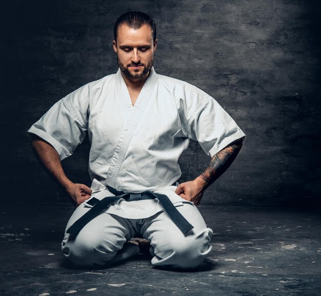 Studio portrait of the bearded brutal karate fighter dressed in a white kimono over grey background.