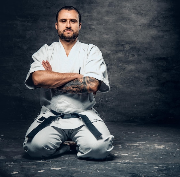 Studio portrait of the bearded brutal karate fighter dressed in a white kimono over grey background.