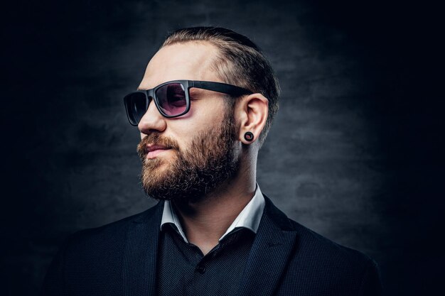 Studio portrait of bearded male dressed in a blue jacket and sunglasses over dark grey background.