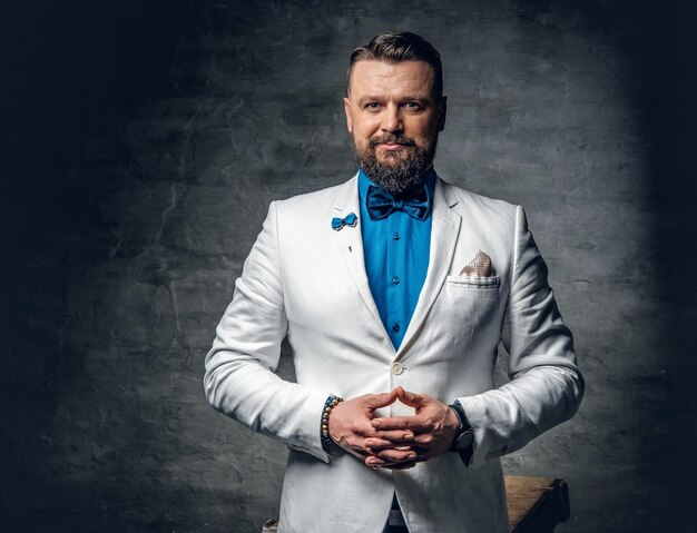 Studio portrait of bearded male dressed in a blue shirt, white jacket and a bow tie over grey background.