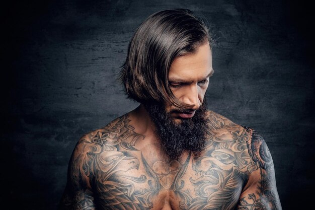 Studio portrait of bearded man with shirtless, tattooed body on grey background.