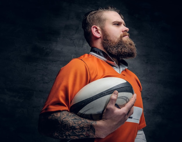 Foto gratuita il ritratto in studio del giocatore di rugby con la barba che indossa una maglietta arancione tiene la palla da gioco.