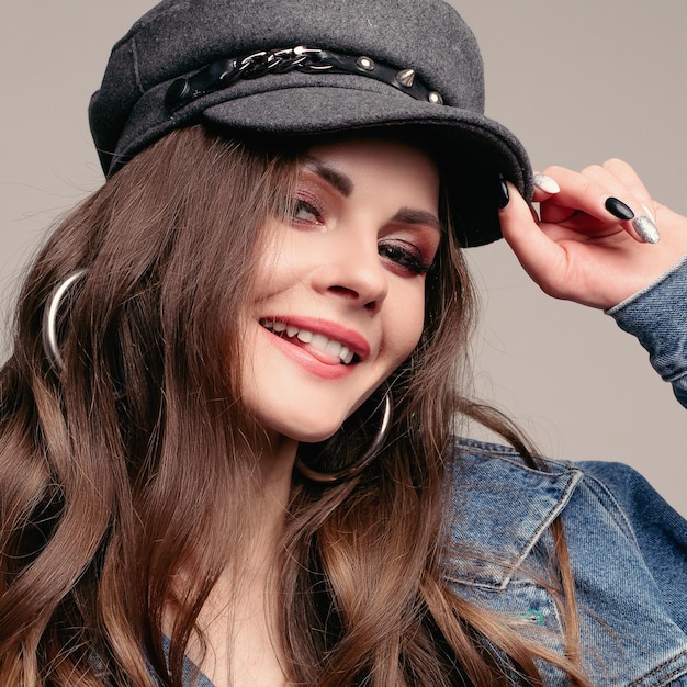 Studio portrait of beautiful brunette Caucasian girl with long wavy hair wearing denim jacket and stylish grey hat She is holding hat with manicured hand and smiling at camera Flirting with camera