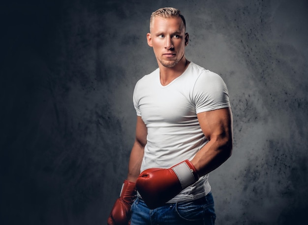 Studio portrait of blond fighter male with red boxing gloves, dresses in a white t shirt over grey background.