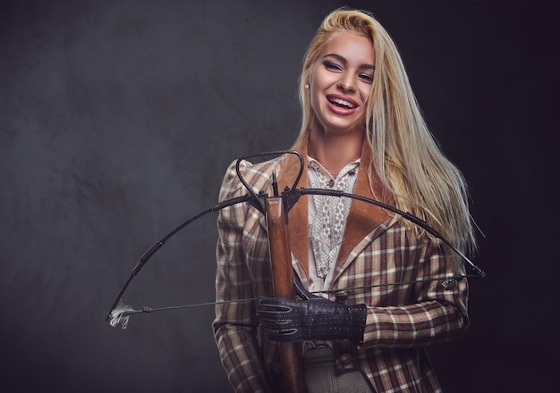 Studio portrait of blond old fashioned female holds a crossbow over grey background.