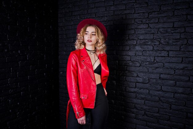 Studio portrait of blonde girl in red hat and leather jacket against brick wall