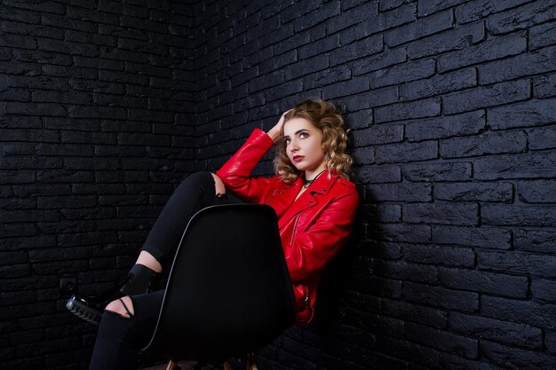 Studio portrait of blonde girl in red leather jacket posed on chair against brick wall