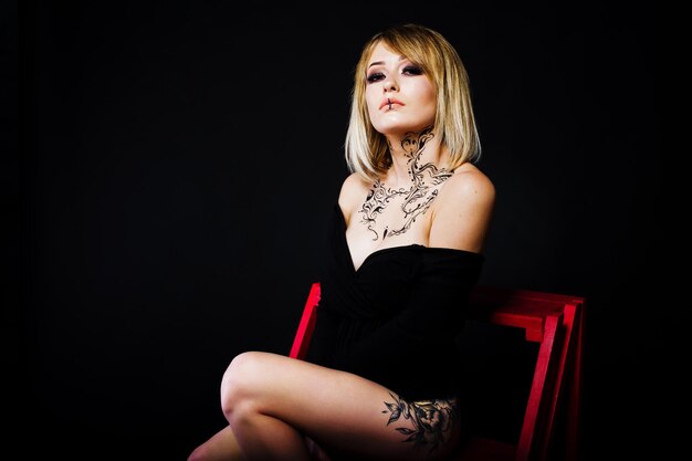 Studio portrait of blonde girl with originally make up on neck and tattoo on thigh wear on black dress at dark background sitting on red ladder