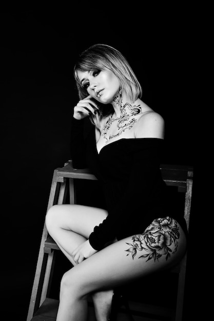 Free photo studio portrait of blonde girl with originally make up on neck and tattoo on thigh wear on black dress at dark background sitting on red ladder