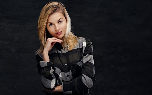 Studio portrait of blonde thoughtful young female dressed in a shirt with square pattern isolated on a dark grey background.