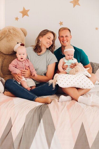 두 명의 유아가 침대에 앉아 있는 쾌활한 가족의 스튜디오 초상화. 플러시 장난감으로 아늑한 침대에 앉아 딸과 아들과 함께 행복한 웃는 어머니와 아버지.