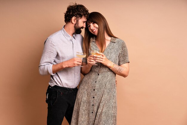 Studio portrait of couple of handsome man and his pretty wife ready for some celebration, holding two glasses of tasty champagne, beige background, elegant outfits, drinking alcohol.