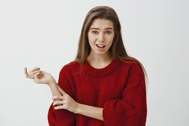 Studio portrait of doubtful unimpressed cute girlfriend in trendy loose sweater, gesturing with head and lifting one eyebrow with disbelief, hearing nonsense, expressing contempt over gray wall