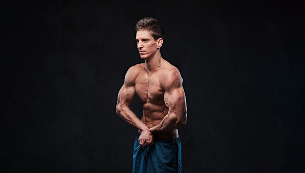 Studio portrait of ectomorph muscle shirtless male shows his biceps over dark grey background.