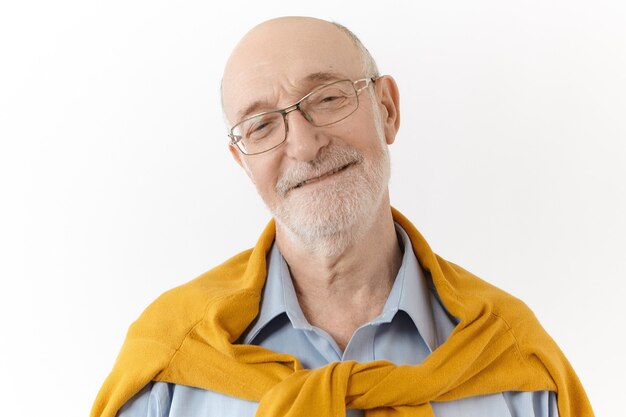 Studio portrait of friendly looking pleased mature seventy year old unshaven grandfather wearing rectangular glasses and stylish sweater over shirt smiling broadly, happy to see his grandchildren