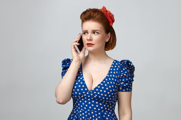 Studio portrait of frustrated beautiful young housewife wearing bright make up and retro dotted dress with low cut having worried facial expression