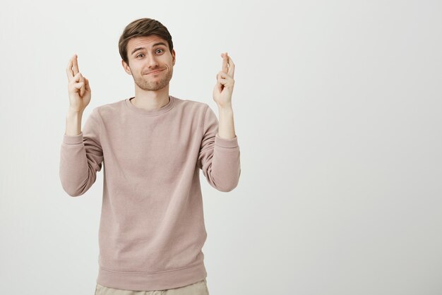 Studio portrait of funny charming male model standing with anxious smile and crossed fingers